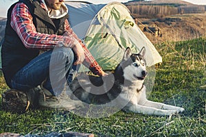 Close-up Guy bearded in jeans checkered shirt and a sleeveless jacket with a dog husk on vacation sitting on nature next