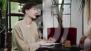 Close-up of a guy sitting in a cafe and writing in a notebook. The waiter girl brings the guy coffee. Coffee house