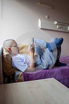 Close up hairless teen girl with phone lying on bed in hospital room, bald female looking at smartphone screen