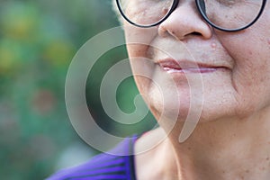 Close-up of half-face a senior woman wearing glasses, smiling and looking at the camera while standing in a garden. Space for text