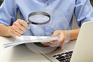 Close up hand of businessperson looking at documents through magnifying glass and laptop in front.