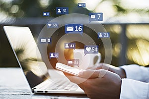 Close up of hand with coffee cup using laptop and smartphone at desktop with social media icons on blurry outdoor background.