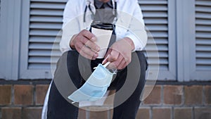 Close-up of hand with cup of coffee and protective medical mask. Tired, exhausted doctor in medical white coat during
