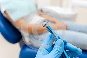 Close-up hand of dentist in the glove holds dental high speed turbine. The patient in blue chair at the background