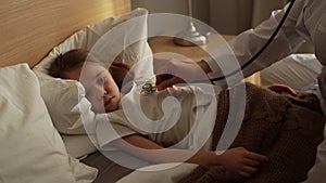 Close-up hand of female doctor listening to heartbeat with stethoscope to check up little sick girl lying in bed.