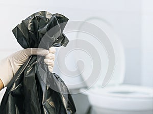 Close up hand with glove holding black garbage bag over blurry white toilet background