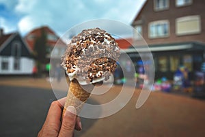 Close up of hand holding cone with white soft serve ice cream covered in chocolate sprinkles with blurry city scene