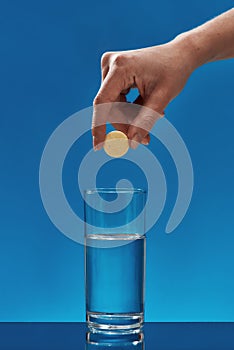 Close up of hand holding effervescent aspirin tablet, pill above glass of water isolated over blue background. Health