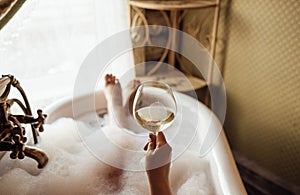 Close-up of a hand holding an elegant glass of champagne in the bathroom. A young woman takes a bubble bath and drinks sparkling