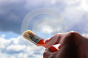 Close-up of hand holding paint brush against sky. Clouds painting. Concept. Sunshine and clouds