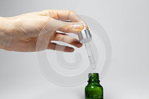 Close-up of hand holding pipette of essential skin care oil, drops in small glass bottle of green. Studio background of grey.