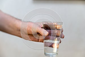 Close-up of a hand holding a tall glass of water against a gray background. A mature man with dirty, untidy fingernails. Glass of