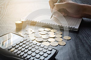 Close up hand of man writing on notebook with calculator with gold coins, pen on black table. Business, finance, marketing, e-