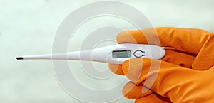 Close up of a hand of the medical worker in orange protective gloves holding white digital thermometer on light blurred background