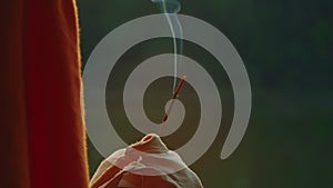 Close-up, the hand of a religious man in orange robes holding a steaming incense and a bag of rosaries