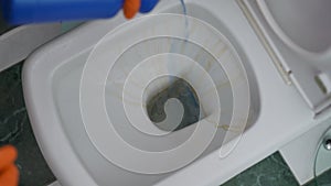 Close-up. A hand in a rubber glove pours blue liquid from a blue plastic bottle into the toilet. The liquid removes