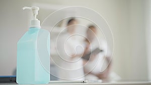 Close-up of hand sanitizer at front with blurred people talking at the background. Doctor consulting patient in hospital