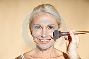 Close up of hand of skillful artist doing natural makeup for mature blonde woman isolated over beige background. The