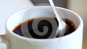 Close-up of Hand stirring sugar in a cup of aromatic coffee with spoon. A cup of aromatic coffee is placed on a wooden table.