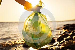 Close up of hand wearing glove and holds bog plastic full trash bag. Ocean in background. Concept of ecological disaster