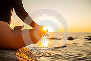 Close up hand of a woman meditating in a yoga lotus pose on the beach at sunset. Girl sitting on a warm rock with sun
