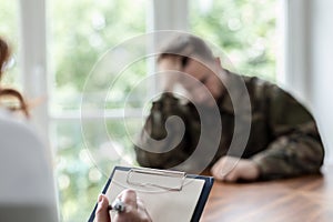 Close-up of a hand writing on a piece of paper with a depressed soldier in the blurred background