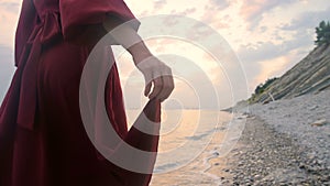 Close-up of the hand of a young girl with a red dress from behind walking on a rocky beach the sea coast at sunset. A