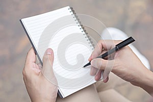 Close up hand young man are using pen writing Record Lecture note pad into the book sitting on the chair outdoor.