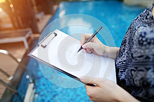 Close-up of hand of young woman signing document on background of sunny day pool. Front used with Open Font License