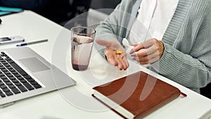 Close up of hands of aged woman, senior intern holding, taking pills out of bottle while using laptop, sitting at desk