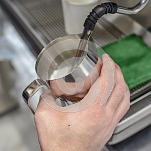 Close-up of hands of a barista frothing warm milk on a coffee maker for making cappuccino or latte in a cafe.