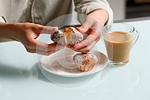 close up of hands breaking apart one of two curd muffins over plate, and glass cup of coffee with milk standing on table.