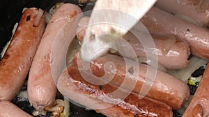 Close up hands chef stirring sausages with metal fork. Stirs and cooking appetizing sausages with onion fried in black
