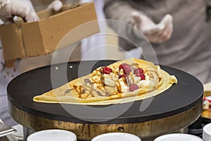 Close-up of hands of cook in gloves preparing golden Crepe, pancake on frying pan with fresh banana, raspberry, sweet