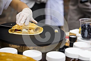 Close-up of hands of cook in gloves preparing rolled up thin Crepe with fresh tasty fruit on frying pan, sweet sauce