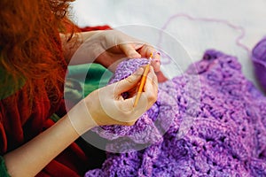 Close-up of hands crocheting. A woman with red hair knits lilac clothes. Selective focus