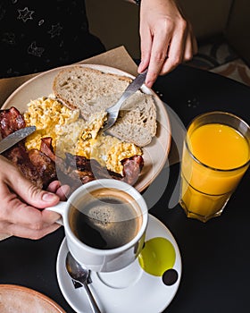 Close-up of hands eating scrambled eggs, bacon, toast, orange juice and coffee