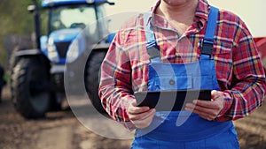 Close-up of hands. Farmer, in red plaid shirt, is typing in tablet smth, against background of tractors, agricultural