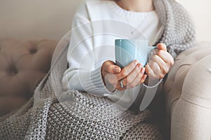 Close up of hands holding blue cup of tea or coffee. Model in white sweater and cozy plaid is sitting, relax at home on sofa