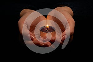 Close-up of hands holding a burning candle in the dark. Adult woman with a candle in her hands on a black background. Selective