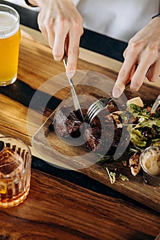 Close up of hands holding knife and fork is cutting grilled ribeye steak on wooden cutting board
