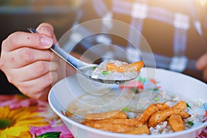 Close up Hands holding spoon with Congee or Rice porridge and deep-fried dough stick, egg ,minced pork balls in bowl