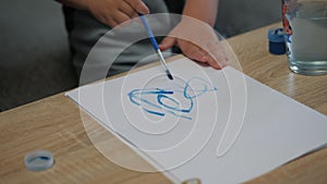 Close-up of hands of little child draws with a brush and blue paint on a sheet of paper at home