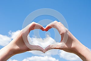 Close-up of hands making a heart symbol on a background of blue sky and clouds. Love, charity, valentines day concept