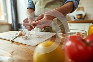 Close up of hands of man in apron peeling garlic while preparing healthy meal, soup in the kitchen. Cooking at home