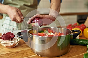 Close up of hands of man, chef cook adding onion to the pot with chopped vegetables while preparing a meal in the