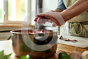 Close up of hands of man, chef cook adding onion to the pot with chopped vegetables while preparing a meal in the