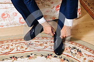 Close-up hands of a man tie laces on black shoes. A businessman in blue trousers and socks wears shoes before going to