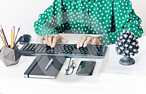 Close-up of hands middle-aged woman in green blouse typing on keyboard laptop computer, concrete holder with pencils and pens,