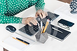 Close-up of hands middle-aged woman in green blouse typing on keyboard laptop computer, concrete holder with pencils and pens,